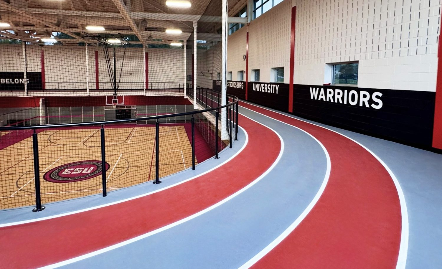 Close-up of the elevated indoor running track curve at ESU's recreation center, with a bold "Warriors" mural on the black wall, the red and gray rubberized track surface in the foreground, and the gymnasium court visible below through the safety railing.