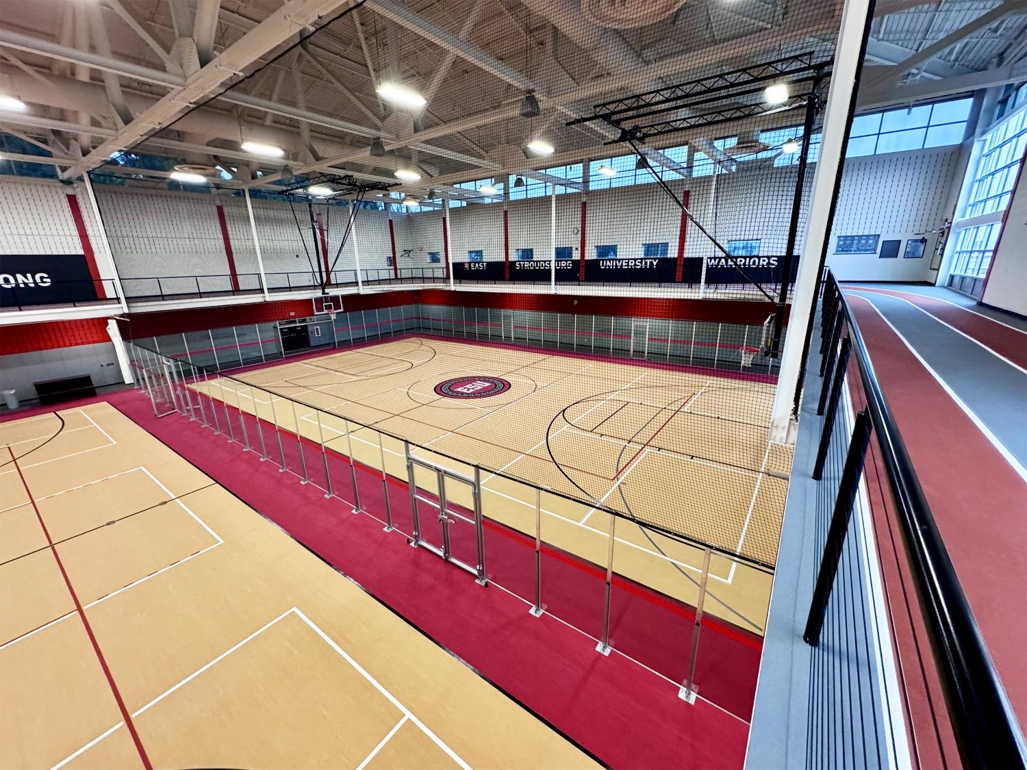 Aerial view from the mezzanine level of East Stroudsburg University's recreation center, showing a hardwood multi-sport floor with basketball court markings, a red running track along the perimeter, and the ESU Warriors logo at center court