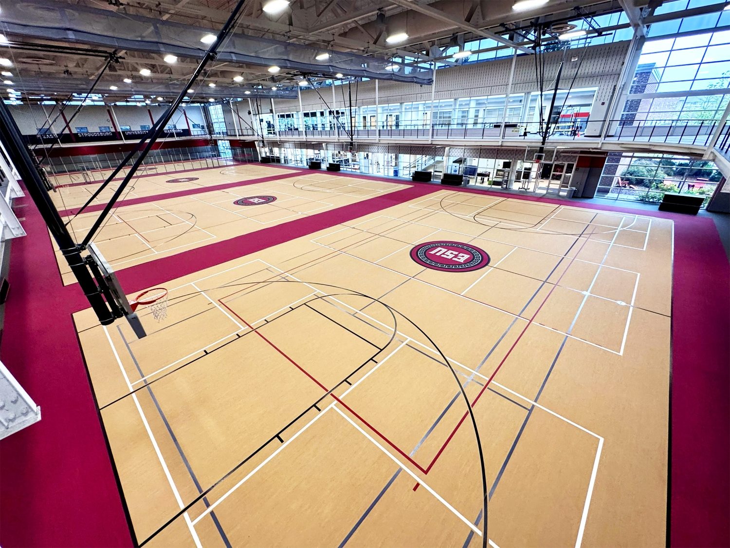 Ground-level wide angle of ESU's recreation center hardwood floor showing multiple overlapping court lines in black, red, gray, and white for basketball, volleyball, and other sports, with the ESU medallion logo visible at center court.