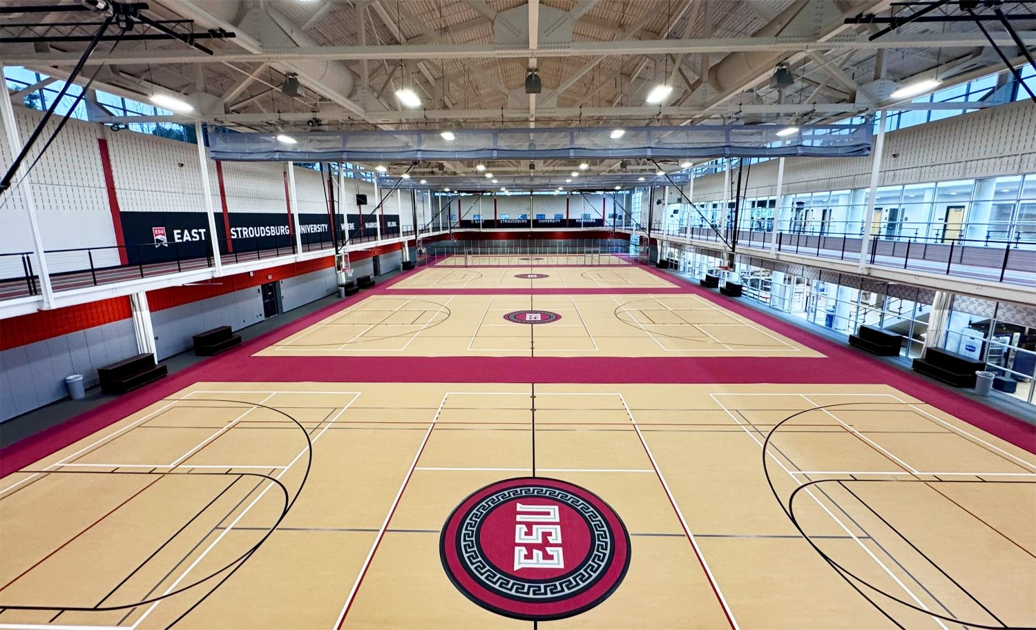 Straight-on view from the upper level of ESU's recreation center, showing the full length of the hardwood gymnasium floor with three court configurations, crimson lane accents, ESU medallion logos, and branded wall banners reading "East Stroudsburg University Warriors."
