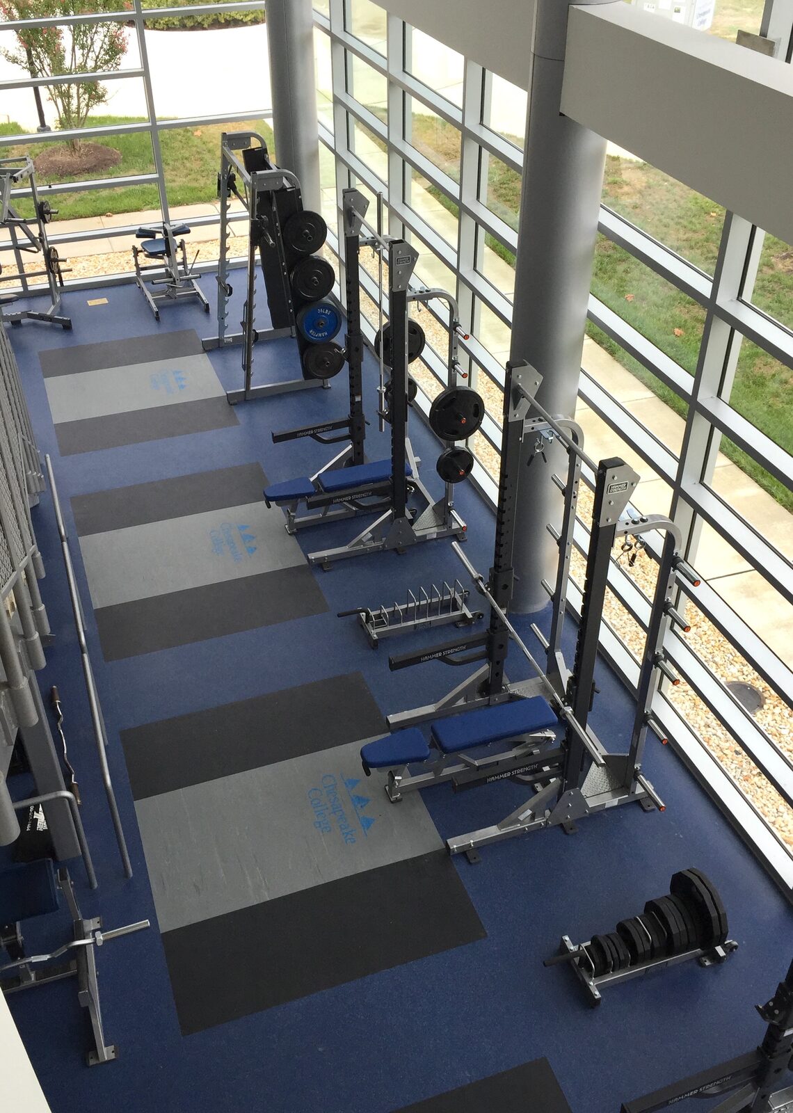 Chesapeake College weight room with a row of Hammer Strength racks and blue and gray flooring featuring the Chesapeake College logo