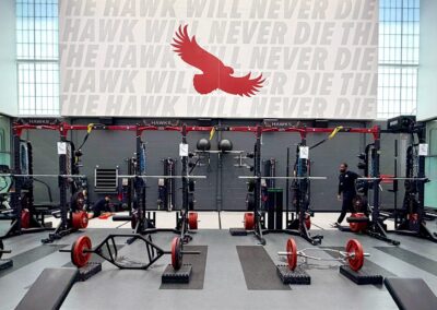 St. Joseph's University weight room close-up with a row of strength racks and hawk logo flooring beneath "The Hawk Will Never Die" wall mural